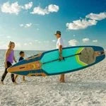 Body Glove Performer 11' inflatable stand-up paddle board on a sandy beach with people.